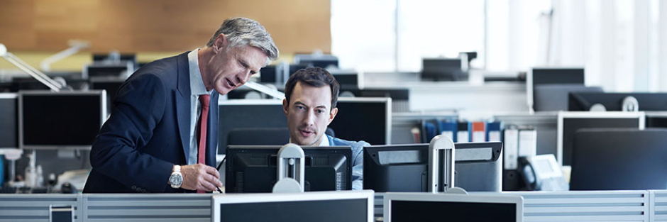 man in suit talking to employee at cubicle desk looking at computer monitors