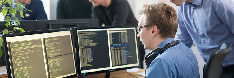 person with glasses looking at computer monitors at office desk with coworkers around