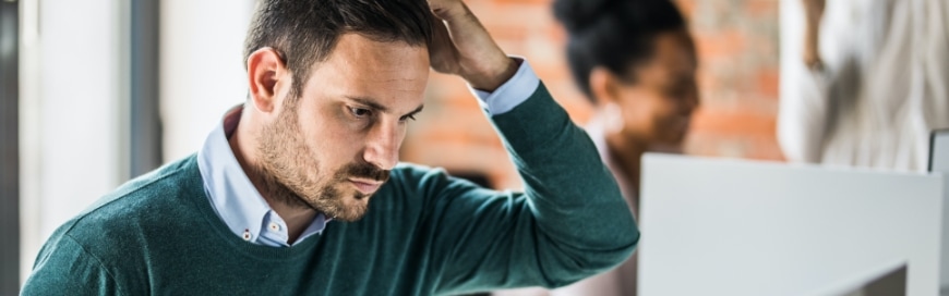 man scratching his head looking confused in an office setting with coworkers in the background